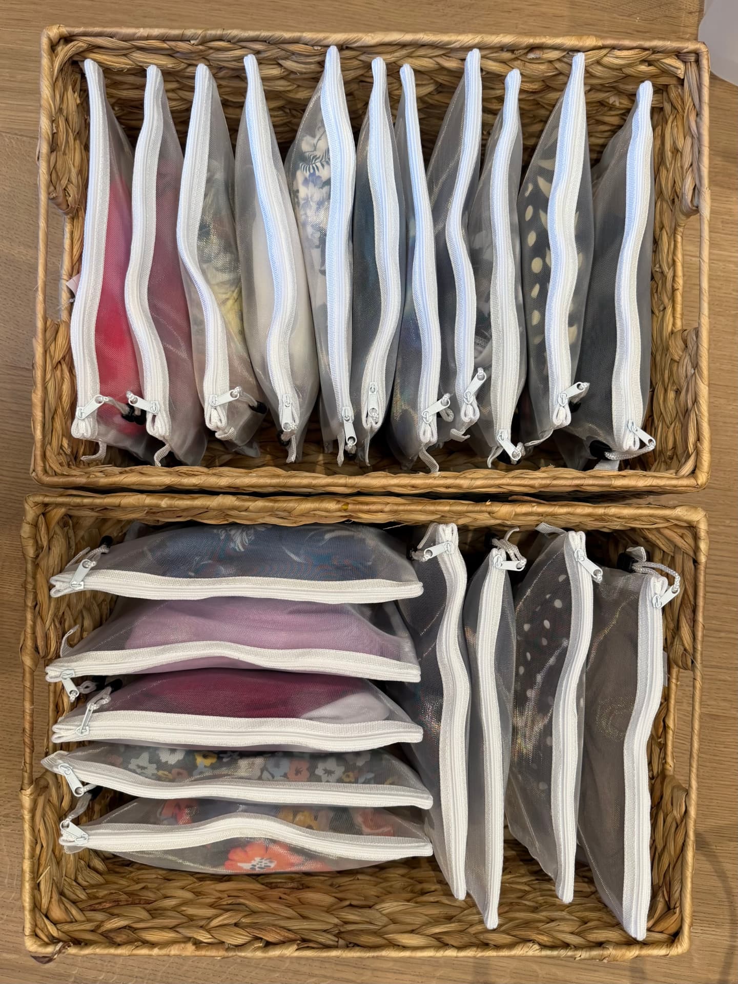 Two wicker baskets holding clear zippered pouches of folded clothes, stored upright and stacked