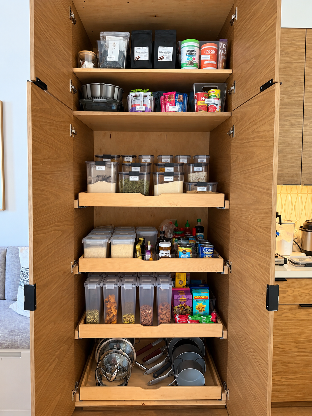 Tall wooden kitchen pantry cabinet with pull-out shelves holding labeled canisters, baskets, and snacks