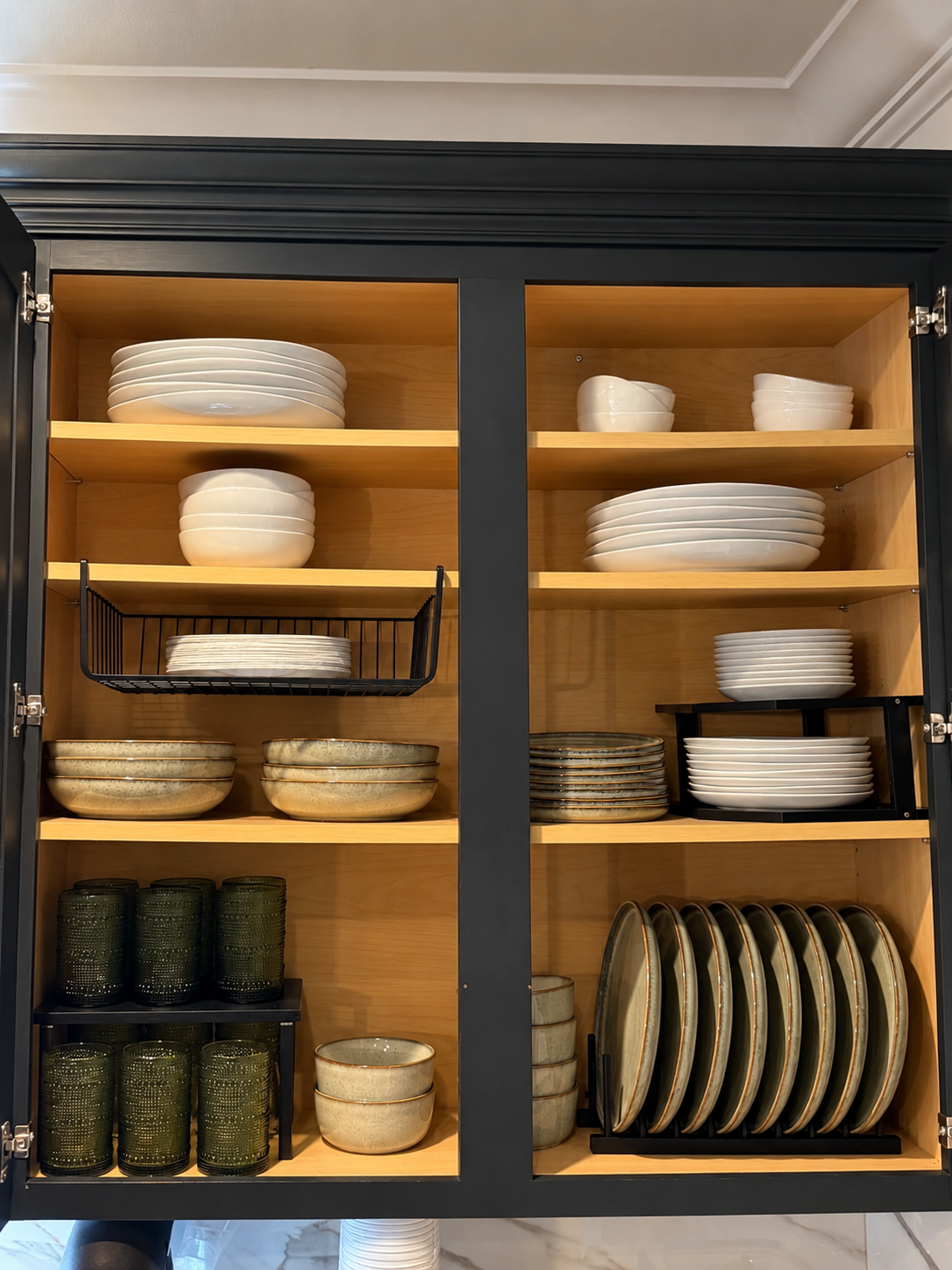 Dark navy cabinet with light wood interior holding stacks of white plates and bowls, sage green plates in a black rack, and dark green glasses