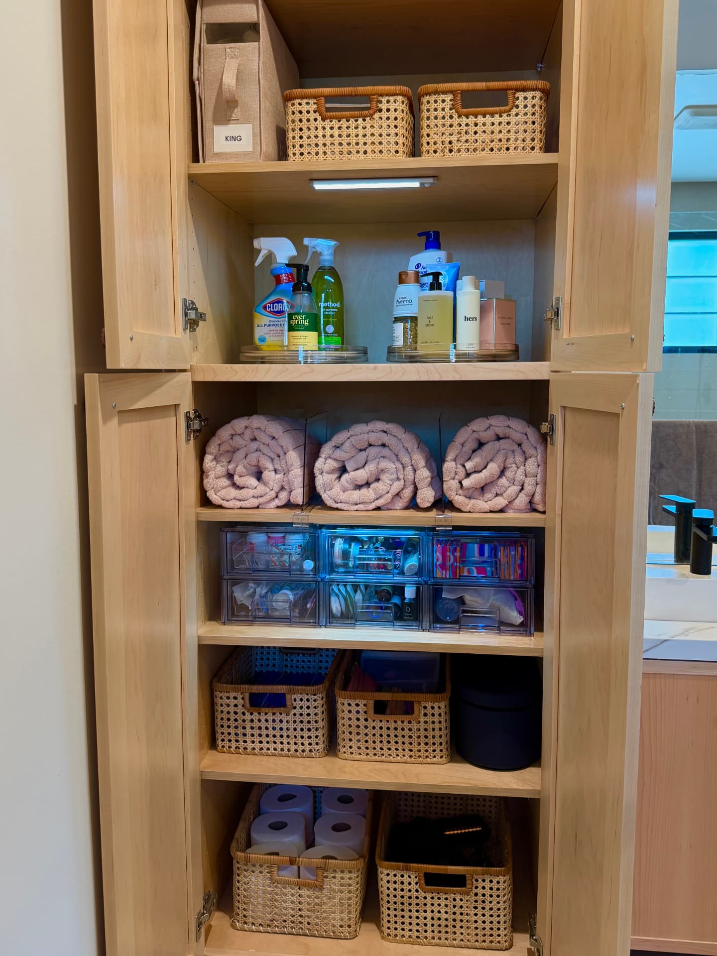 Full bathroom cabinet with a labeled king bin, cane baskets, clear turntables of bath and cleaning products, rolled pink towels divided with clear acrylic, and stacked clear drawers