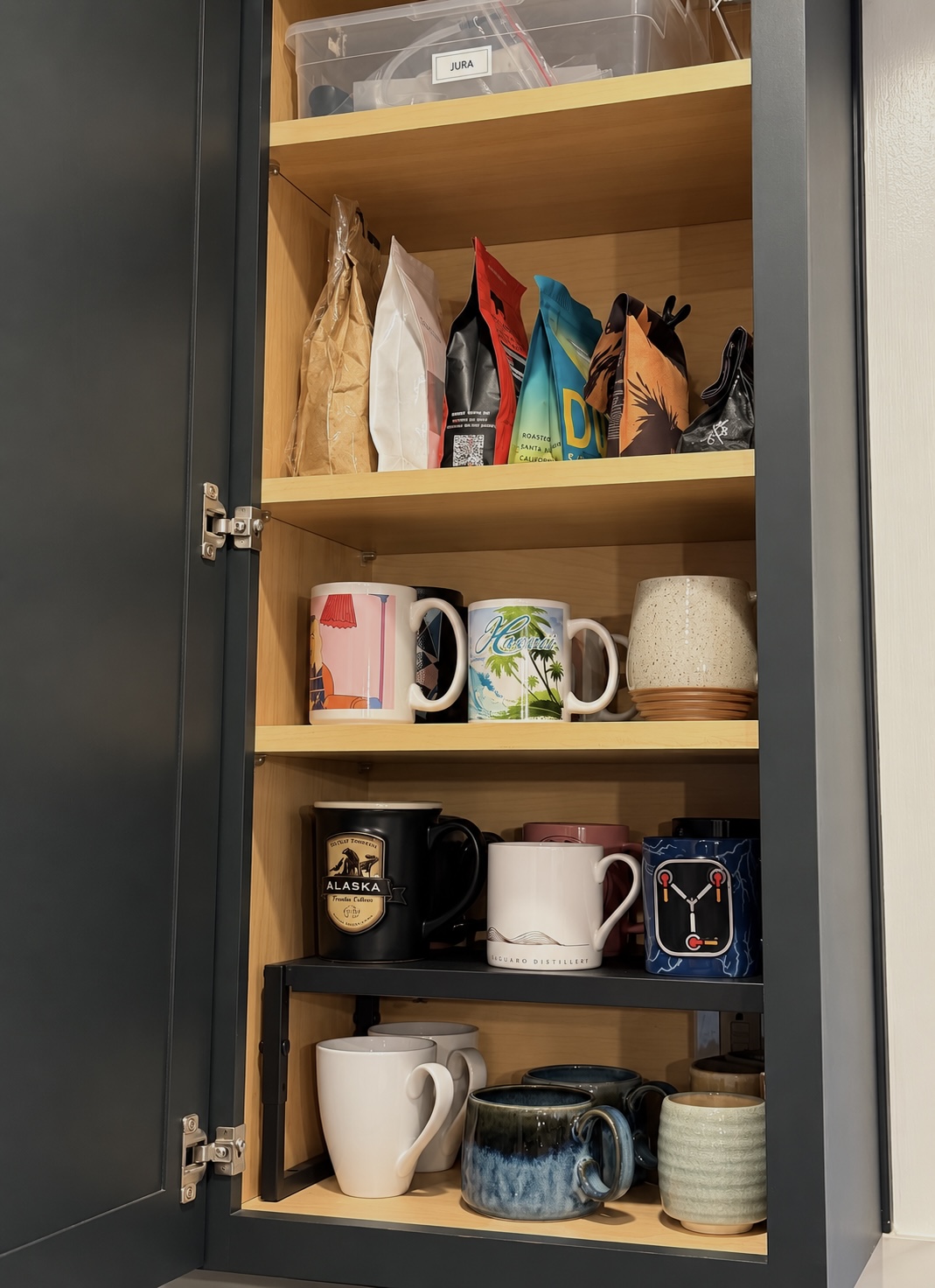 Upper kitchen cabinet coffee station with bags of coffee beans on one shelf and rows of mugs on a black metal riser below