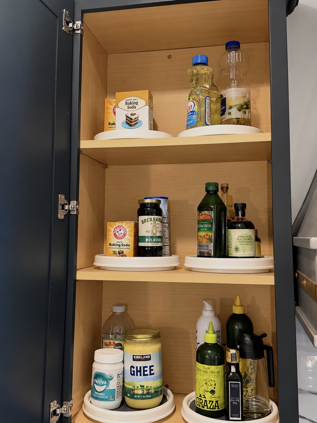 Three pantry shelves with white turntables holding baking soda, molasses, olive oils, and ghee